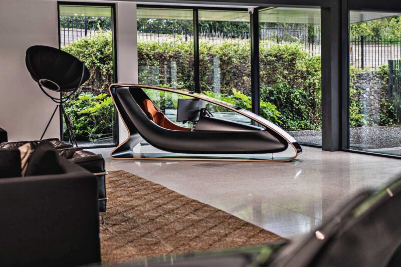 A futuristic, black ergonomic workstation inspired by the Collector’s Racing Sim—designed by an ex-Jag designer and Prodrive with B&O sound—features a curved frame and built-in monitor, positioned by large glass windows in a modern, bright room.