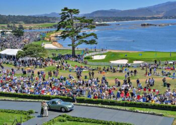 A large crowd gathered on a grassy area near the ocean with mountains in the background, watching a classic green car on display at an outdoor event, reminiscent of the Pebble Beach Concours set for an earlier date in 2028.