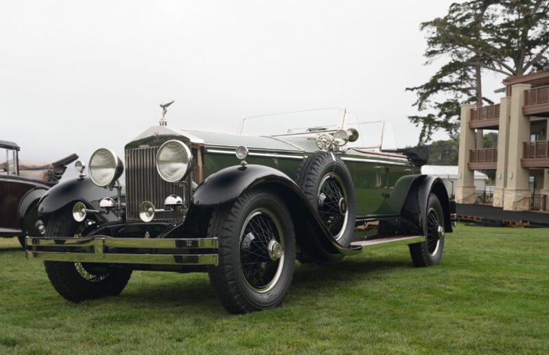 A vintage green Rolls-Royce Phantom convertible with black fenders is parked on grass near buildings and trees, capturing the timeless elegance of Pebble Beach over 100 years.