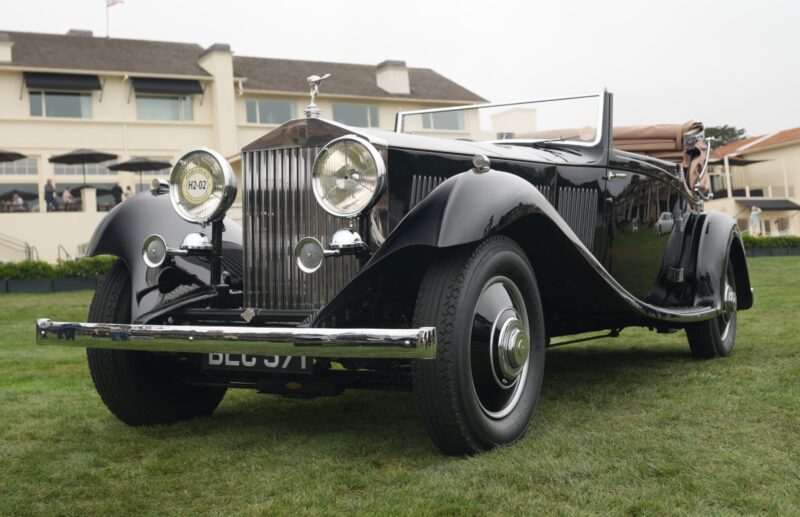 A classic black Rolls-Royce Phantom convertible is parked on grass in front of a large building at Pebble Beach, viewed from a low front angle, capturing over 100 years of motoring elegance.