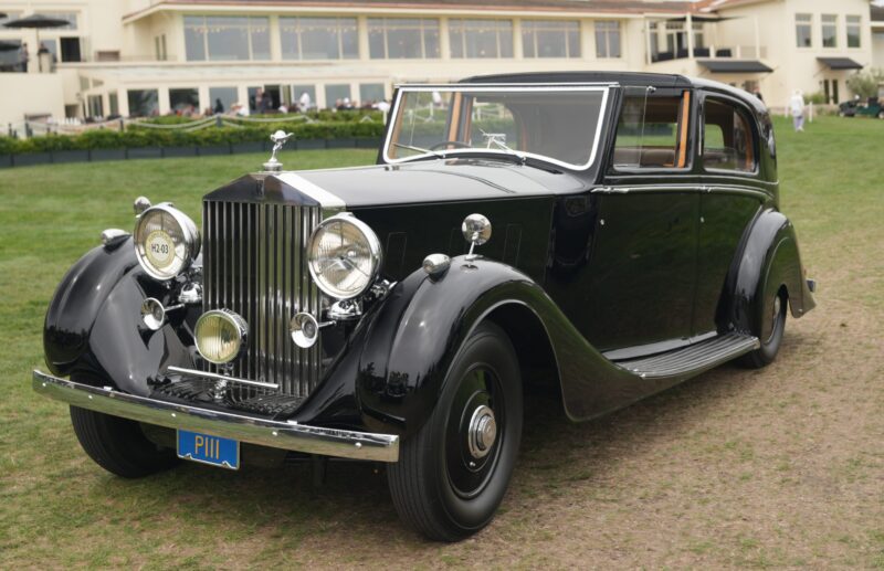 A classic black Rolls-Royce Phantom is parked on grass in front of a large building with multiple windows, celebrating 100 Years of elegance at Pebble Beach.