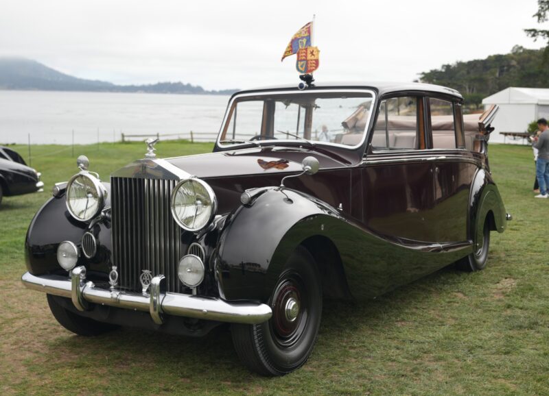 A classic black and maroon Rolls-Royce Phantom limousine is parked on grass, with a hood ornament and flag on the roof, overlooking a scenic waterfront—capturing timeless elegance at Pebble Beach’s 100 Years celebration.