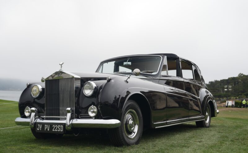 A classic black Rolls-Royce Phantom sedan is parked on grass at Pebble Beach, with a misty background and distant trees, celebrating 100 Years of luxury.