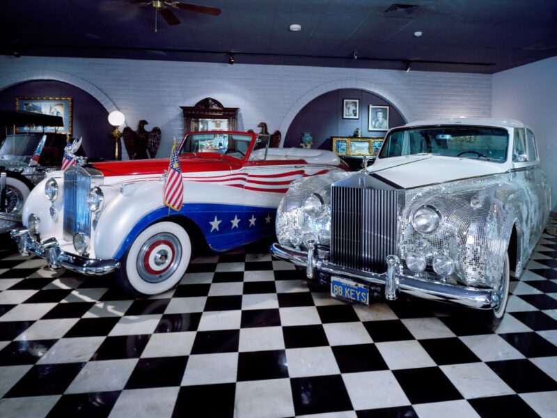 Two vintage Rolls-Royce cars—one with an American flag motif, the other shimmering with mirror-like tiles—are displayed indoors on a black and white checkered floor, evoking the glamorous spirit of music and luxury.