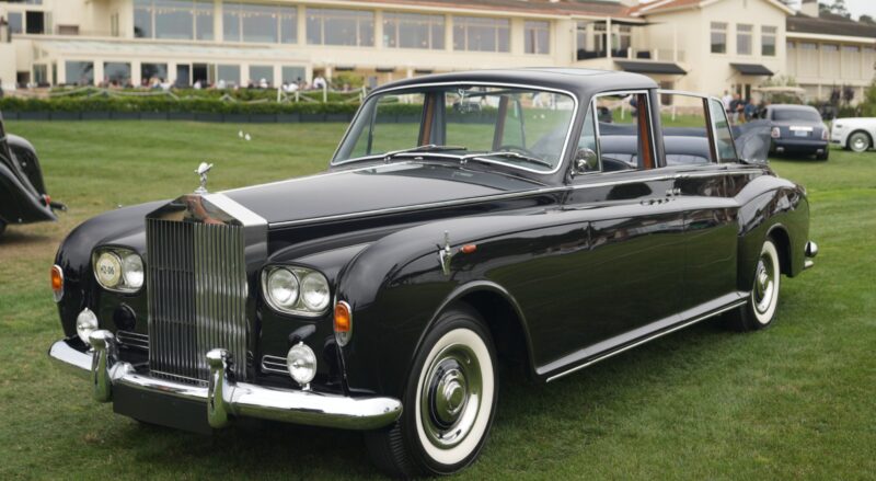 A classic black Rolls-Royce Phantom sedan is parked on grass in front of a large building at Pebble Beach, with people visible in the background.