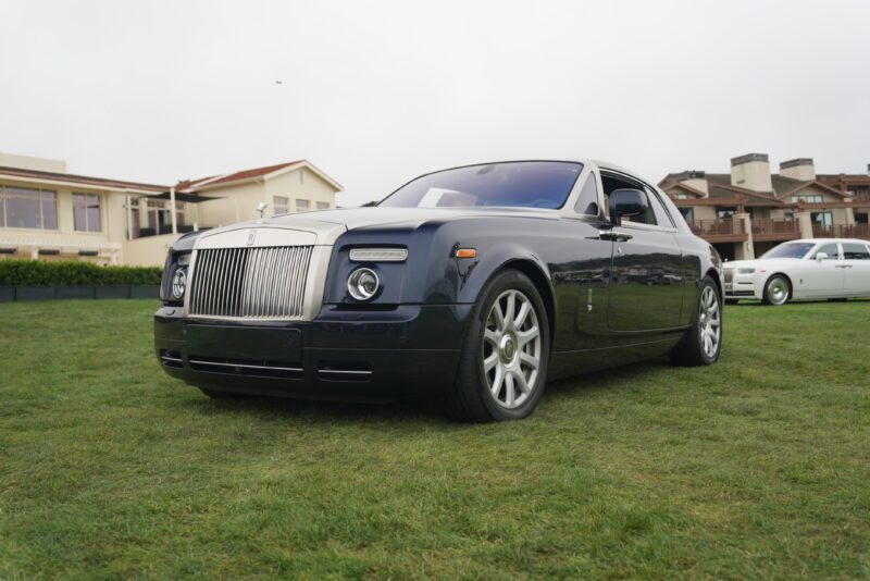 A black Rolls-Royce Phantom coupe, celebrating 100 Years of luxury, parked on grass with residential buildings in the background.