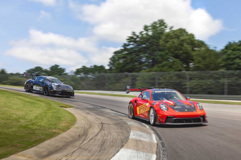Two race cars speed around a curve on the racetrack, the red car leading closely followed by the black; trees and sky in the background. The Porsche 911 GT3 RS Manthey Package is now offered at Porsche Track Experience in Alabama.