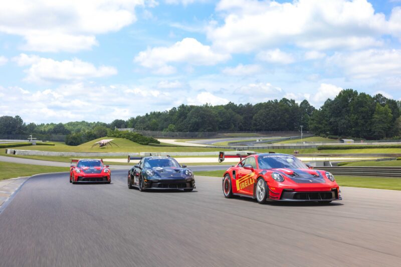 Three sports cars, including one with Pirelli branding, race side by side on a track surrounded by green hills and trees, as the Porsche 911 GT3 RS Manthey Package is now offered at the Porsche Track Experience in Alabama under a partly cloudy sky.