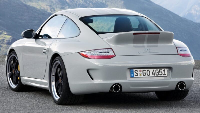 A silver Porsche 911 sports car, perfect for a Porsche 911 Comparison: 997 Sport Classic Vs 992 Sport Classic, is parked outdoors with mountains in the background, viewed from the rear left side.
