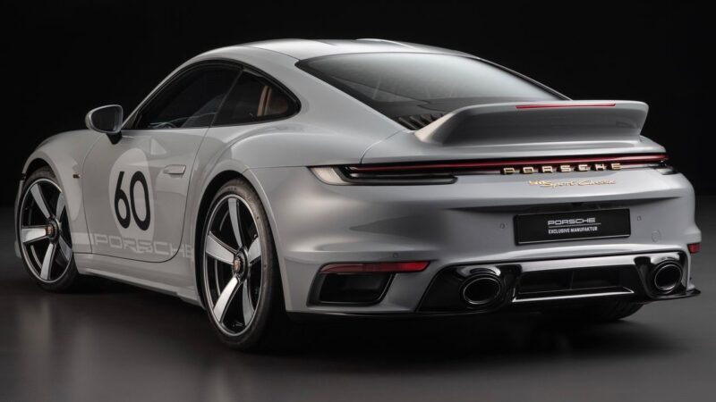A silver Porsche 911, showcasing the number 60 on its door and a raised rear spoiler, is parked in a studio with a dark background—perfect for a Porsche 911 comparison: 997 Sport Classic vs 992 Sport Classic.