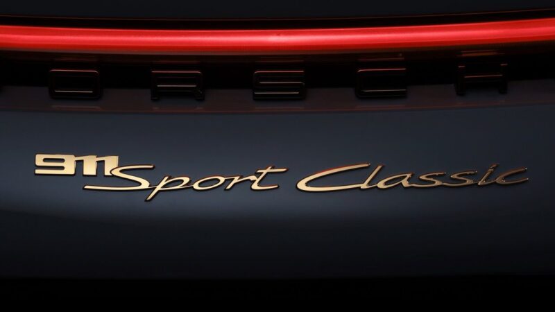 Close-up of the rear badge on a Porsche car, showing "911 Sport Classic" in gold lettering with part of a red taillight above—a striking detail for any Porsche 911 Comparison: 997 Sport Classic vs 992 Sport Classic.