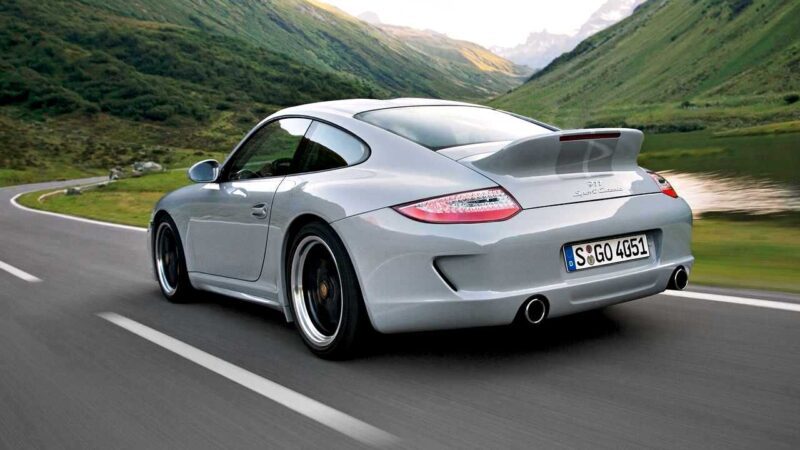 A silver Porsche sports car drives on a winding road through green mountains beside a river, showcasing the thrill that comes alive in every Porsche 911 Comparison: 997 Sport Classic vs 992 Sport Classic.