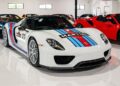 A white Porsche 918 Spyder with Martini Racing livery, one of the most expensive Porsches of all time, is parked indoors among other sports cars.