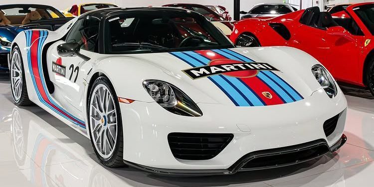 A white Porsche 918 Spyder with Martini Racing livery, one of the most expensive Porsches of all time, is parked indoors among other sports cars.