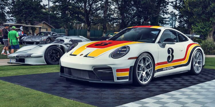 A white Porsche 911 GT3 ANDIAL Wrap with red and yellow racing stripes and the number 3 on the hood is displayed outdoors beside another silver sports car at a show where Penske’s 963 RSP headlines Monterey.