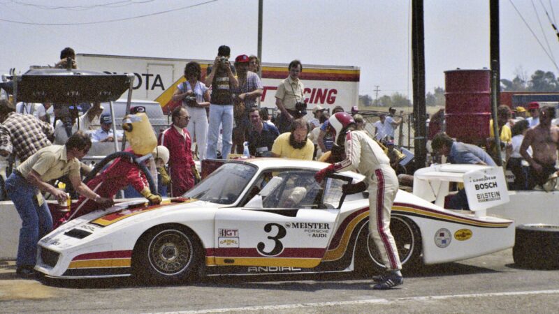 A pit crew services a white Porsche 911 GT3 ANDIAL Wrap race car with the number 3 on the side during a pit stop at a motorsport event, while spectators and team members watch.