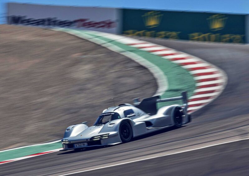 A silver prototype race car, reminiscent of the Porsche 911 GT3 ANDIAL Wrap, speeds around a curving racetrack with red, white, and green curbs under a clear sky.