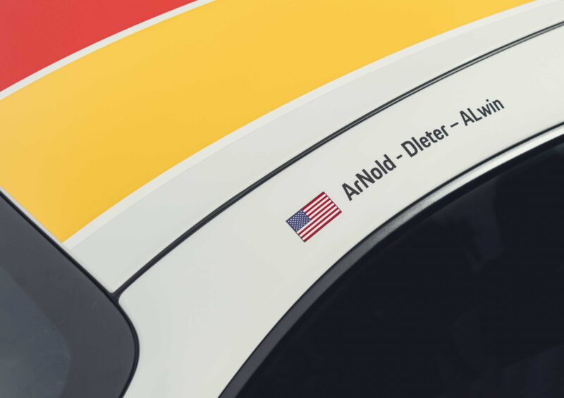Close-up of a car roof with a red and yellow stripe, an American flag decal, and the names "Arnold-Dieter-Alwin" beside it, inspired by the iconic Porsche 911 GT3 ANDIAL Wrap.
