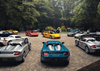 Several Porsche Carrera GT sports cars, part of a 2026 Porsche Plática event, are parked in a circular pattern on a stone courtyard, surrounded by trees and people in the background.