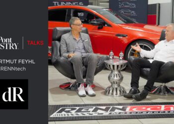 Two men in conversation sit in chairs with a table between them, a red AMG car in the background, and duPont REGISTRY and RENNtech branding displayed.