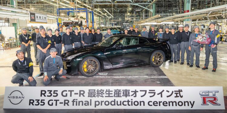 A group of factory workers poses with the last Nissan R35 GT-R beside a sign reading "R35 GT-R final production ceremony," marking the end of an era inside an automotive assembly plant.
