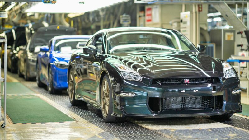 A black Nissan R35 GT-R leads a line of vehicles on an automotive assembly line inside a factory, with machinery and equipment visible in the background—capturing the end of an era and hinting at what's next.