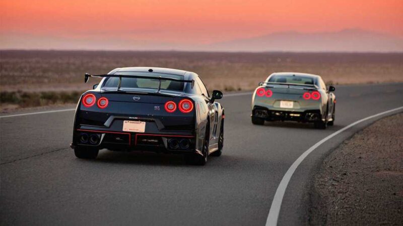 Two sports cars, including a Nissan R35 GT-R, speed along a curved desert road at sunset. Mountains loom in the distance under an orange sky, capturing the thrill of what's next at the end of an era.