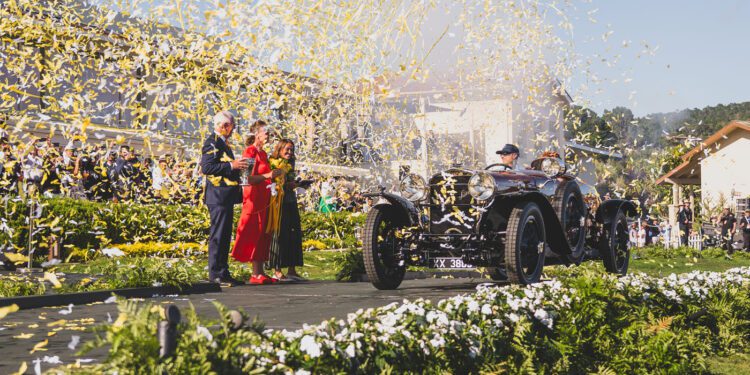 A vintage Hispano-Suiza drives on stage surrounded by yellow confetti, as people applaud nearby at the Pebble Beach Concours d’Elegance after winning Best of Show at this prestigious outdoor event.