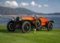 A vintage Hispano-Suiza with a wooden body and exposed wheels is parked on grass near a body of water with hills in the background; a trophy sits in front, evoking the prestige of Pebble Beach Concours d’Elegance.