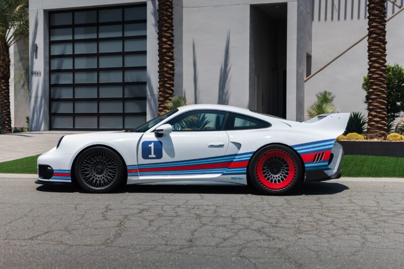 A white Porsche 935 sports car with Martini Racing-style blue and red stripes and number 1 on the door is parked on a street in front of a modern building and palm trees—a true dream come true for any car enthusiast.