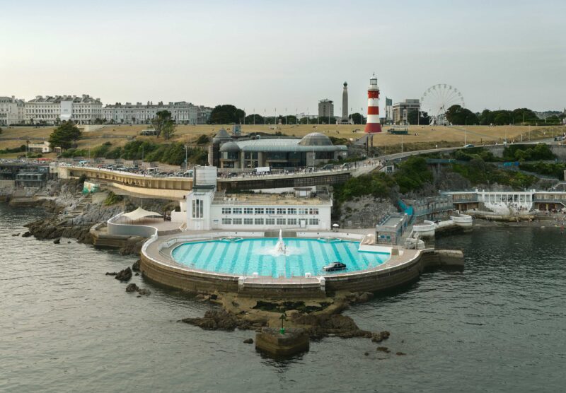 A large circular outdoor swimming pool sits beside the sea, with buildings, a lighthouse, and a Ferris wheel visible on the grassy hill in the background—a setting worthy of a Phantom or the smooth rhythms of music drifting in the breeze.