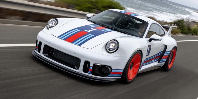 A white Porsche 935 with blue and red racing stripes and red wheels, the ultimate dream car, drives on a scenic coastal road.