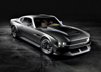 A sleek, modern silver sports car with a retro design evokes the spirit of the Ringbrothers Debut Aston Martin OCTAVIA: a supercharged 1971 DBS reimagined in carbon fiber, parked on a dark, reflective floor in a dimly lit setting.