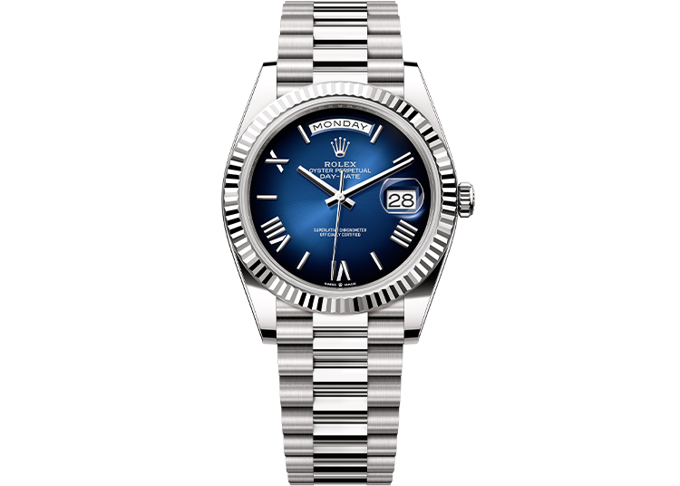 A silver Rolex watch with a blue dial, displaying the day at 12 o'clock and date at 3, featuring a fluted bezel and metal bracelet—a timeless choice for watch enthusiasts during Monterey Car Week.