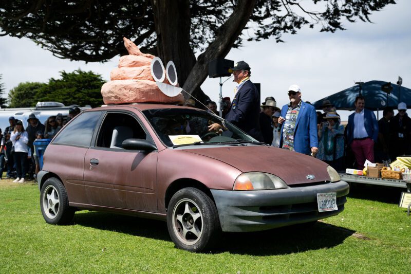 A small, worn red car with a large poop emoji sculpture on its roof is parked on grass at an outdoor event during the 2025 Monterey Car Week, as people from duPont REGISTRY and other attendees stand nearby observing.