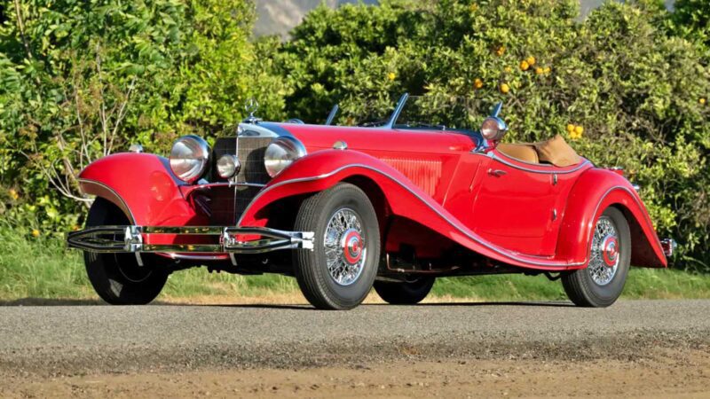 A classic red vintage convertible car with chrome accents is parked on a paved road, surrounded by lush green trees—reminiscent of the 10 Most Expensive Cars To Sell At 2025 Monterey Car Week Auctions.