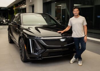 A man stands next to a black Cadillac SUV inside a modern showroom, resting his right hand on the car's hood and smiling at the camera.