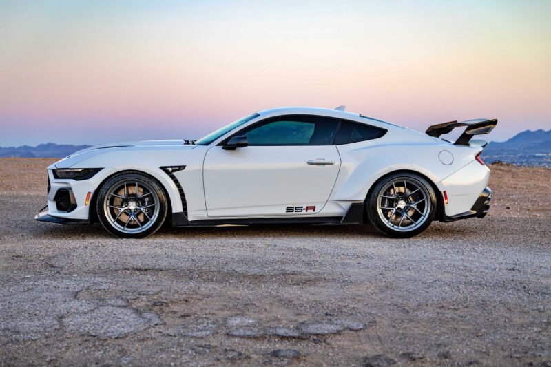 A white Shelby American Debuts 2026 Wide Body Super Snake-R, an 850+ HP track weapon with coil suspension and a rear wing, is parked on gravel with mountains and a pastel sky in the background.