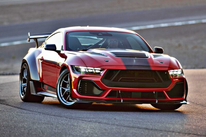 A red sports car with black accents and a large rear spoiler, inspired by the Shelby American Debuts 2026 Wide Body Super Snake-R, races on a paved track, its powerful 850+ HP engine blurring the terrain in the background.