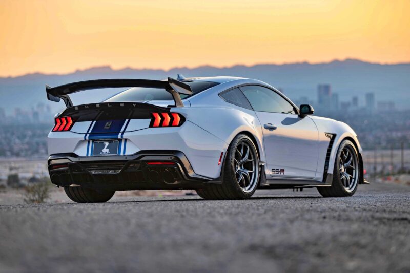 A Shelby American Debuts 2026 Wide Body Super Snake-R, an 850+ HP track weapon with coil suspension, sits parked on a gravel surface at sunset, its white exterior with blue racing stripes framed by a city skyline and mountain backdrop.