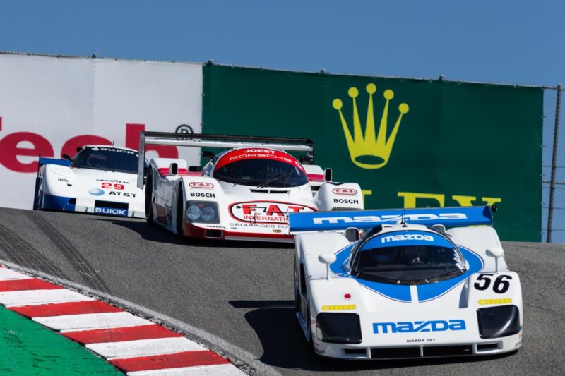 Three racing cars, led by a white and blue Mazda with number 56, navigate a turn on the racetrack during Monterey Car Week 2025, with duPont REGISTRY banners visible in the background.