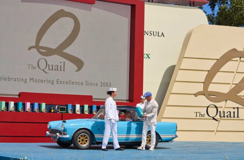 Two men in vintage attire stand beside a blue classic car on stage at The Quail, where the Ferrari F50 GT1 was crowned Best of Show at The Quail A Motorsports Gathering 2025, with trophies visible in the background.