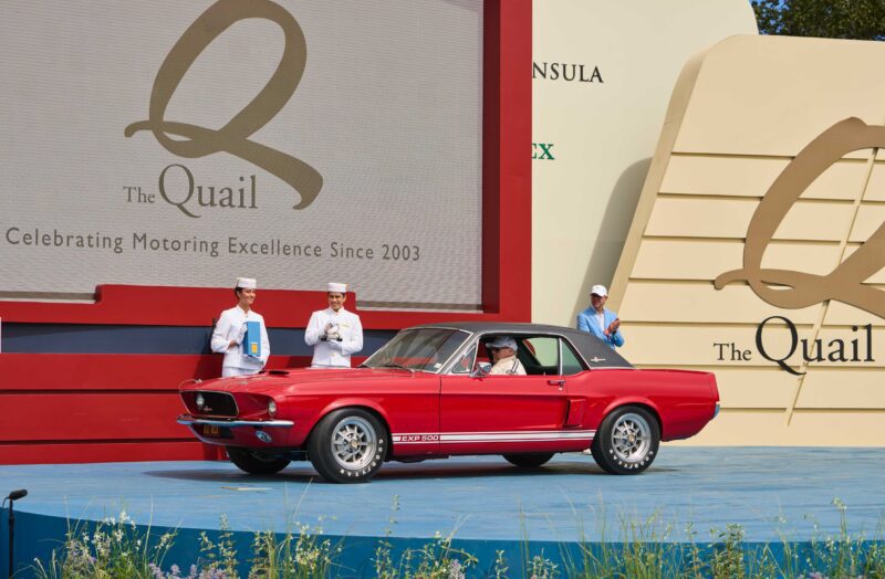 A red vintage Ford Mustang is displayed on an outdoor stage at The Quail, where attendants in white uniforms stand nearby. The event also celebrated the Ferrari F50 GT1 crowned Best of Show at The Quail A Motorsports Gathering 2025.