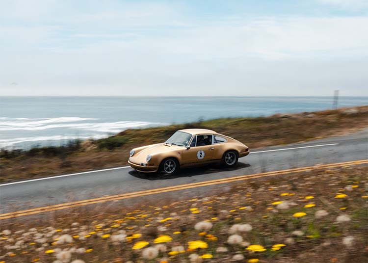 A vintage tan sports car cruises along a coastal road, the ocean and wildflowers in the background—an unforgettable scene reminiscent of 2025 Monterey Car Week and duPont REGISTRY showcases.