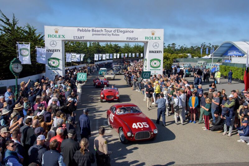 Classic cars drive past large crowds at the starting line of the Pebble Beach Tour d’Elegance, a highlight of Monterey Car Week, with banners, sponsor logos, and luxury watches on display overhead.
