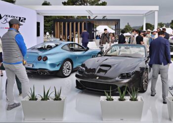 Two luxury sports cars are displayed on a white platform at an outdoor event, as several people admire the Touring Superleggera Debuts New Veloce12 “Barchetta” Restomod Based On The Ferrari 550 Maranello.