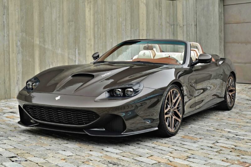 A sleek black Touring Superleggera Debuts New Veloce12 “Barchetta” Restomod, based on the Ferrari 550 Maranello, with a tan interior is parked on a cobblestone surface in front of a concrete wall.