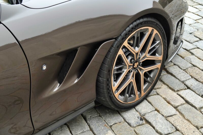 Close-up of the front wheel and side air vent of a luxury sports car, inspired by the Touring Superleggera Veloce12 “Barchetta” restomod based on the Ferrari 550 Maranello, parked on a cobblestone surface.