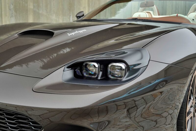Close-up of the front left headlight and hood of a sleek, dark-colored sports car with "Superleggera" script, reminiscent of the Touring Superleggera Debuts New Veloce12 “Barchetta” Restomod Based On The Ferrari 550 Maranello.