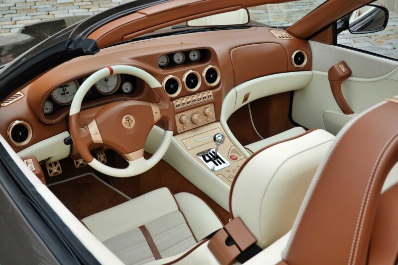 The interior of a luxury convertible car, inspired by the Touring Superleggera Debuts New Veloce12 “Barchetta” Restomod Based On The Ferrari 550 Maranello, features tan and cream leather seats, a matching dashboard, round air vents, and a manual gear shift.